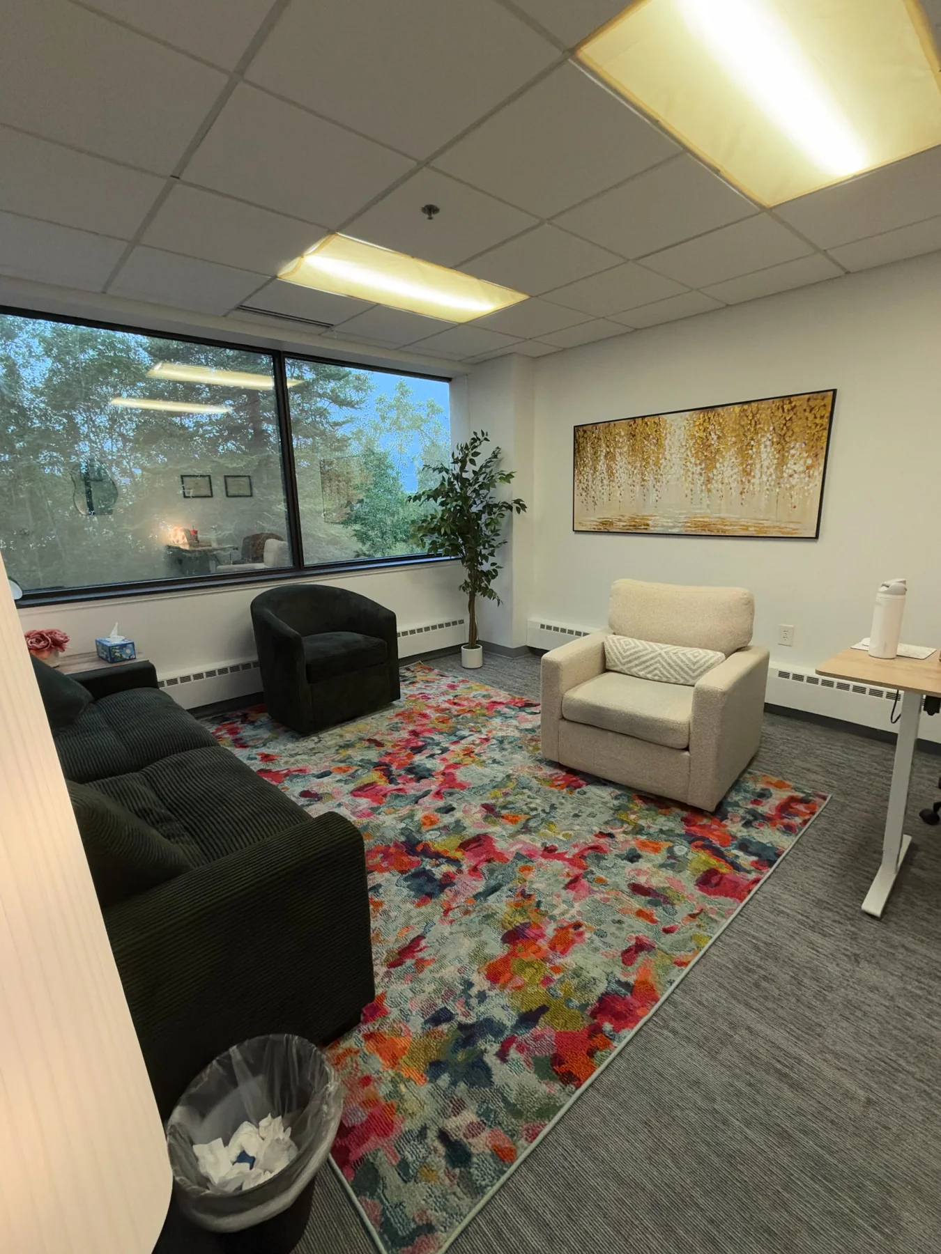 Therapy room with a colorful rug, dark sofa, cream chair, large windows, and gold wall art
