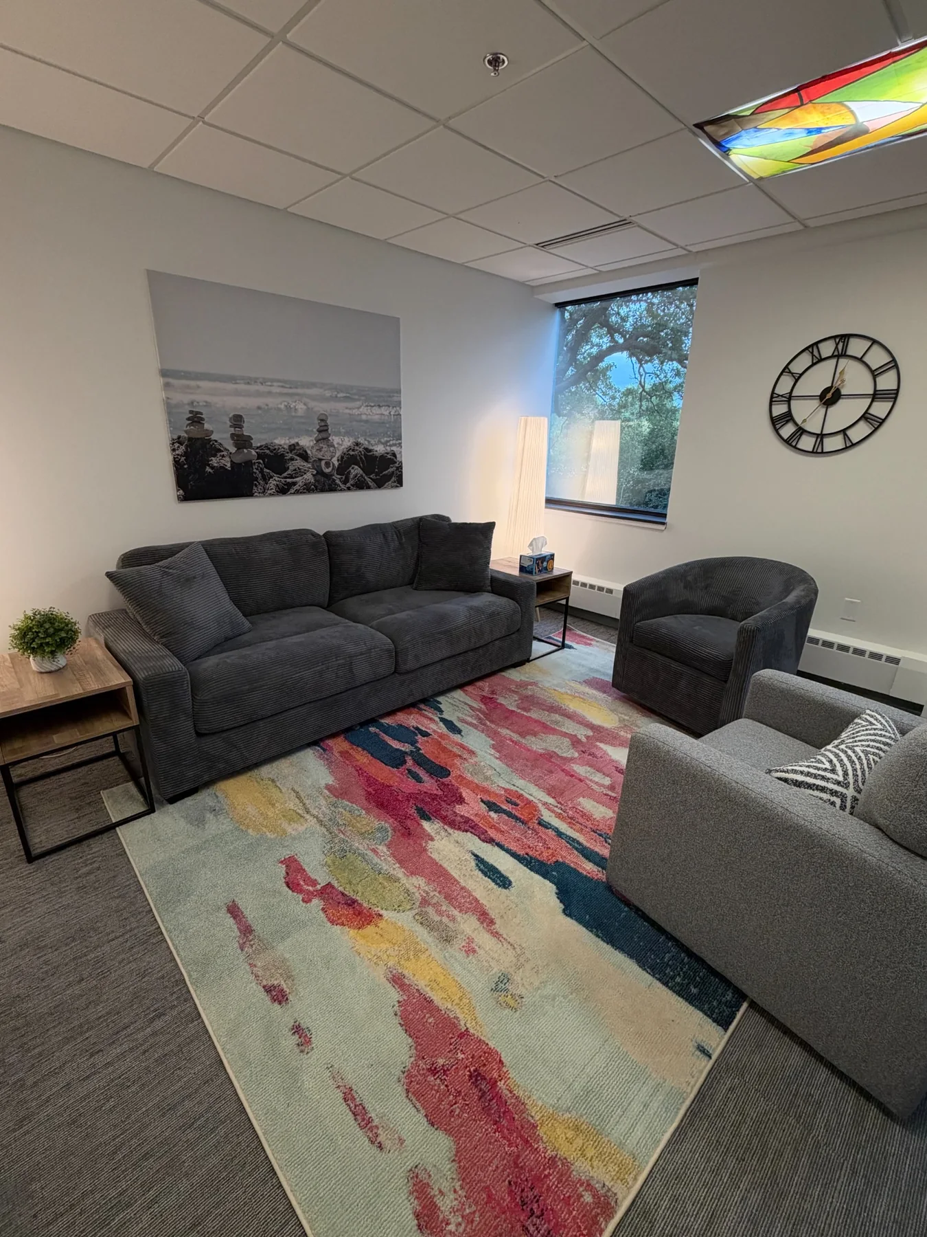 Therapy room with a dark sofa, lounge chairs, colorful rug, and wall art